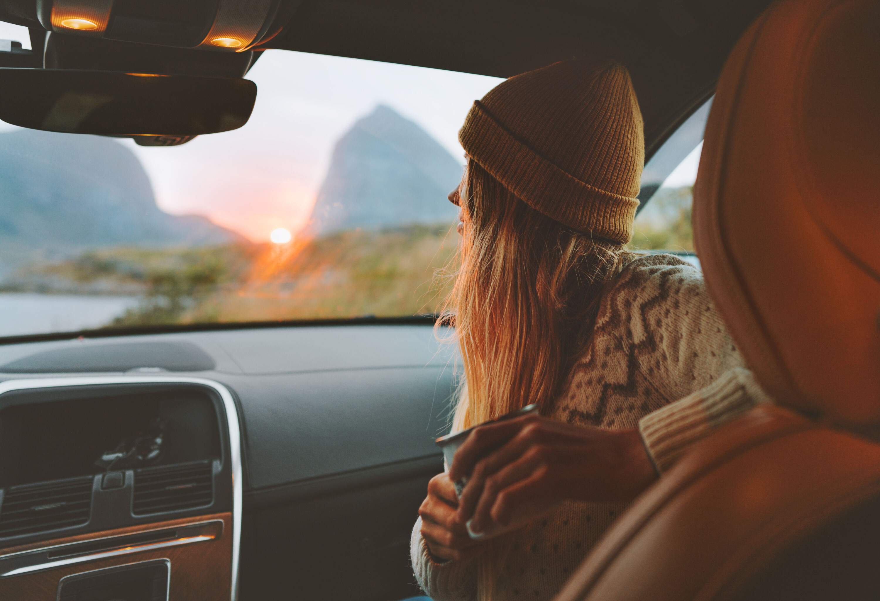 A long-haired woman with a cap sitting in the passenger seat of a car, clutching a cup of coffee.