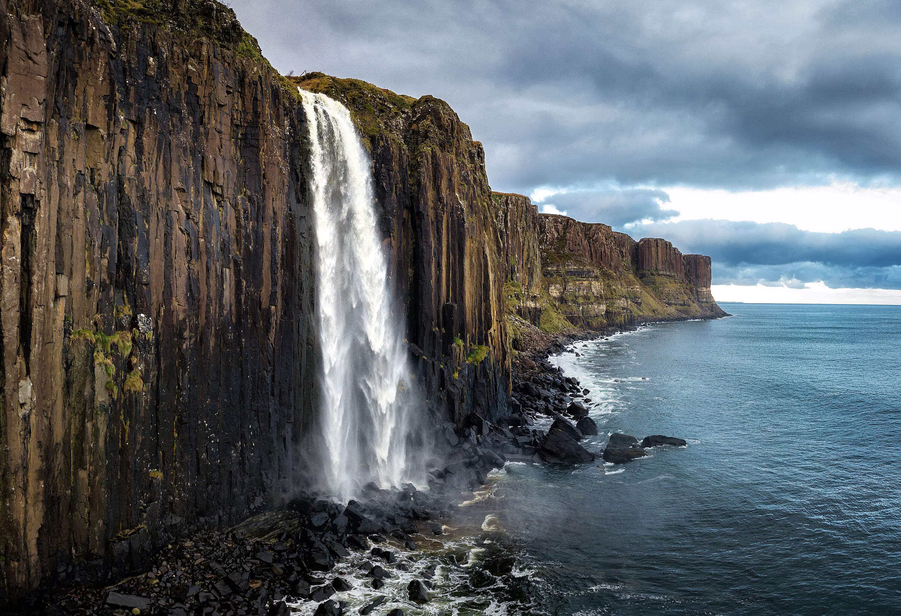 An amazing waterfall plunges into the sea on a rock cliff along a series of vertical basalt columns.