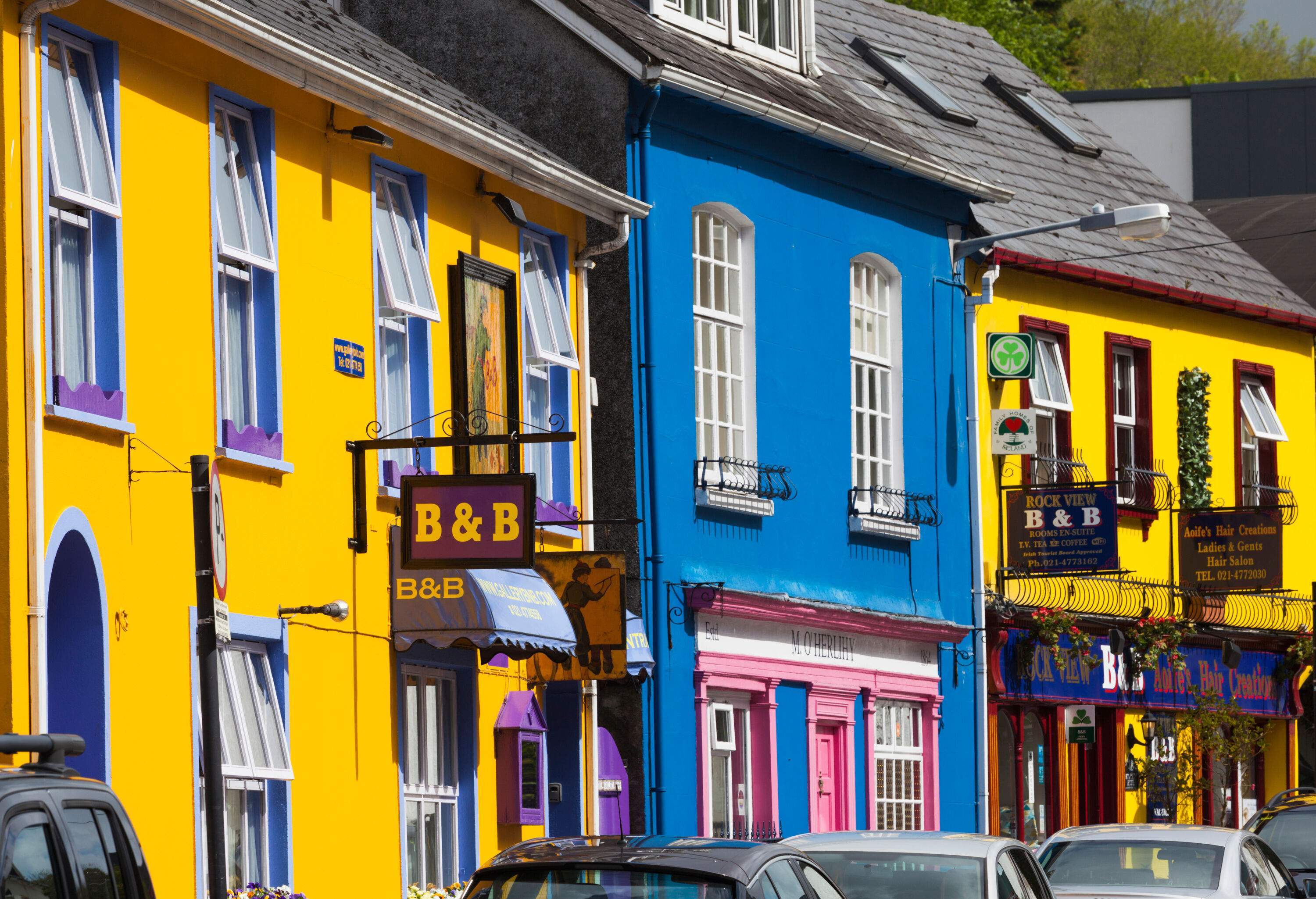 A sunlit row of colourful buildings lined with parked cars.