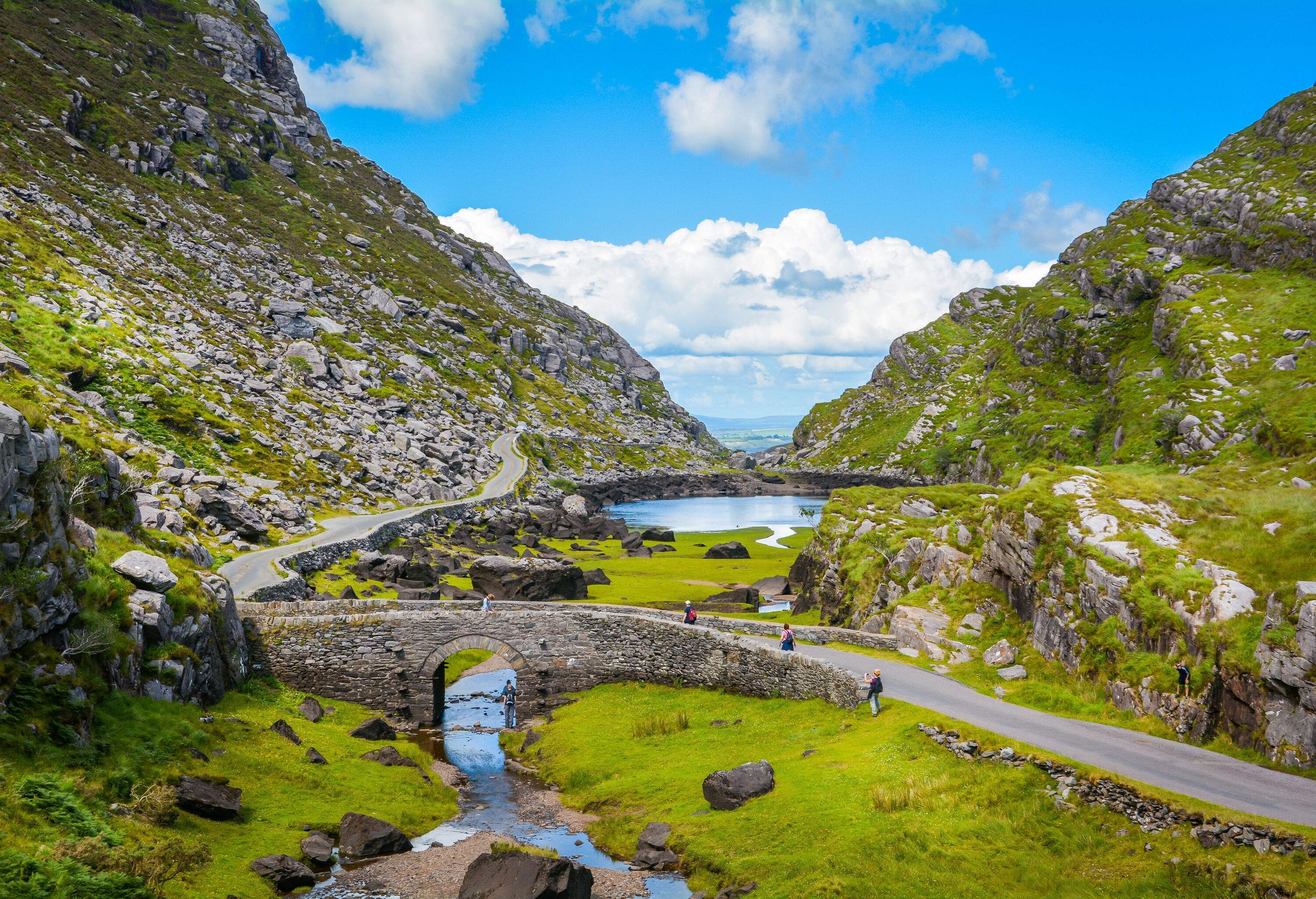 People cross a brook on a stone bridge set in a valley nestled among rocky slopes.