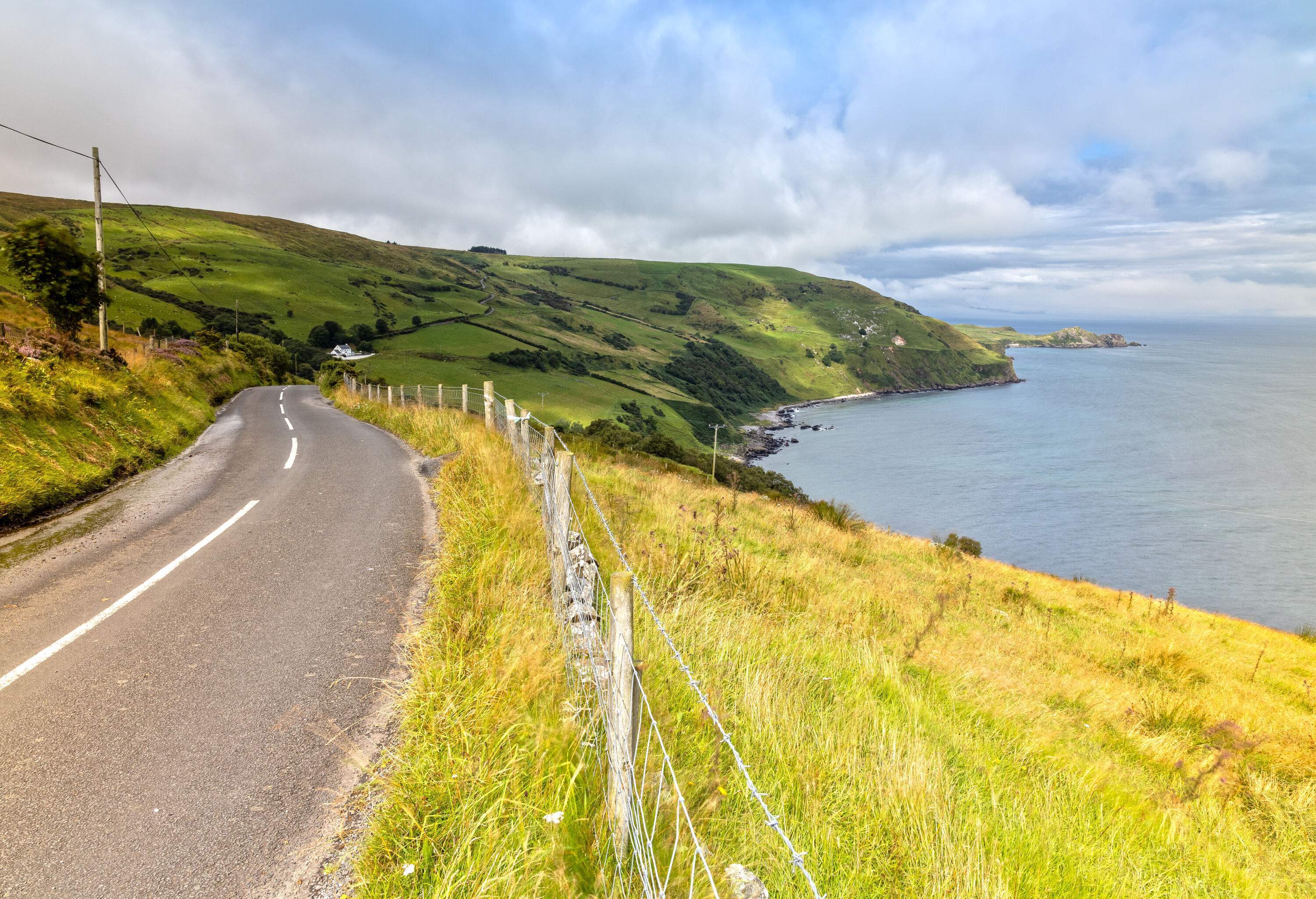 A hillside roadway by the sea with a barbed-wire fence along the grass field.