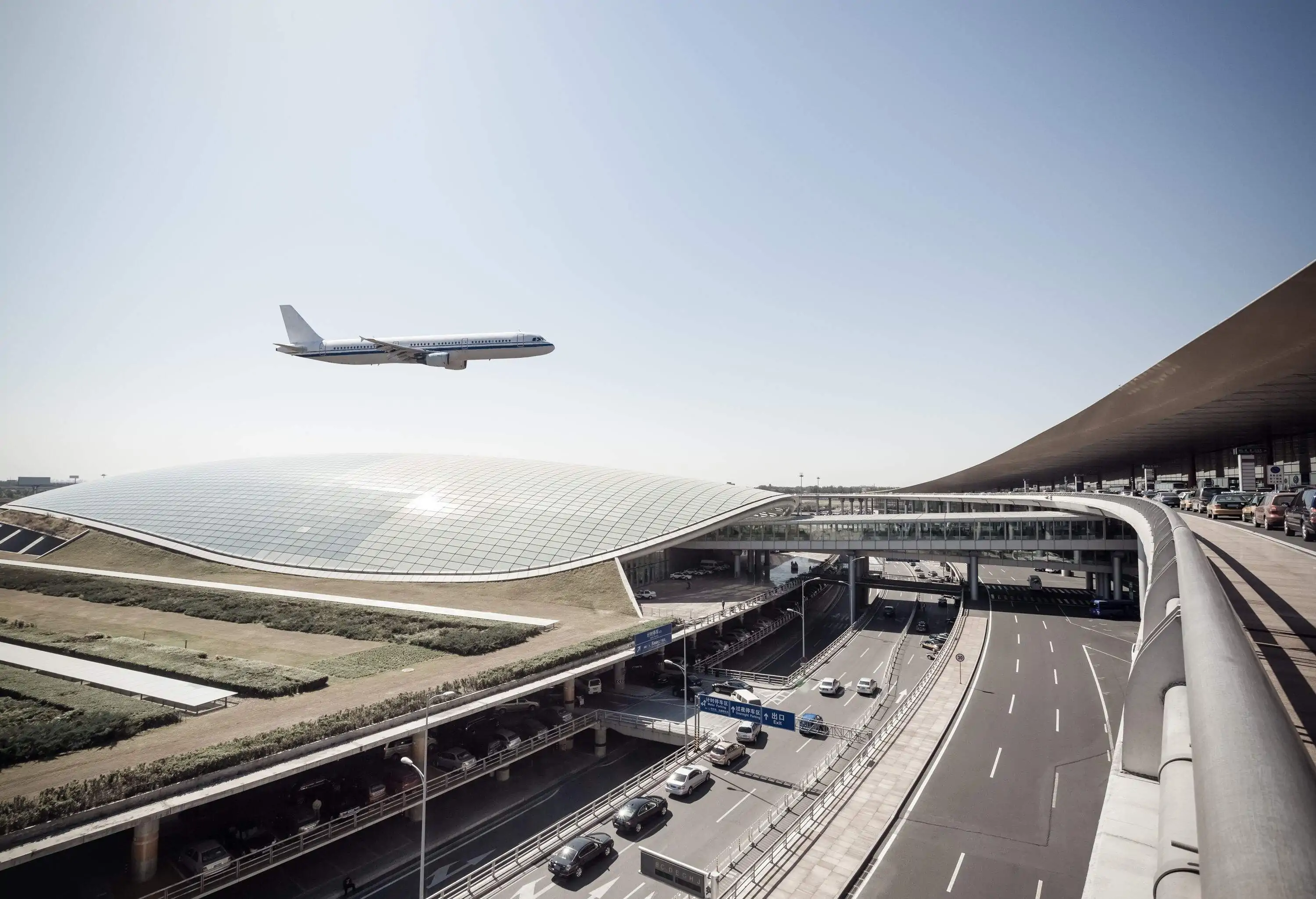 An aeroplane over a futuristic airport with a curve glass roof.