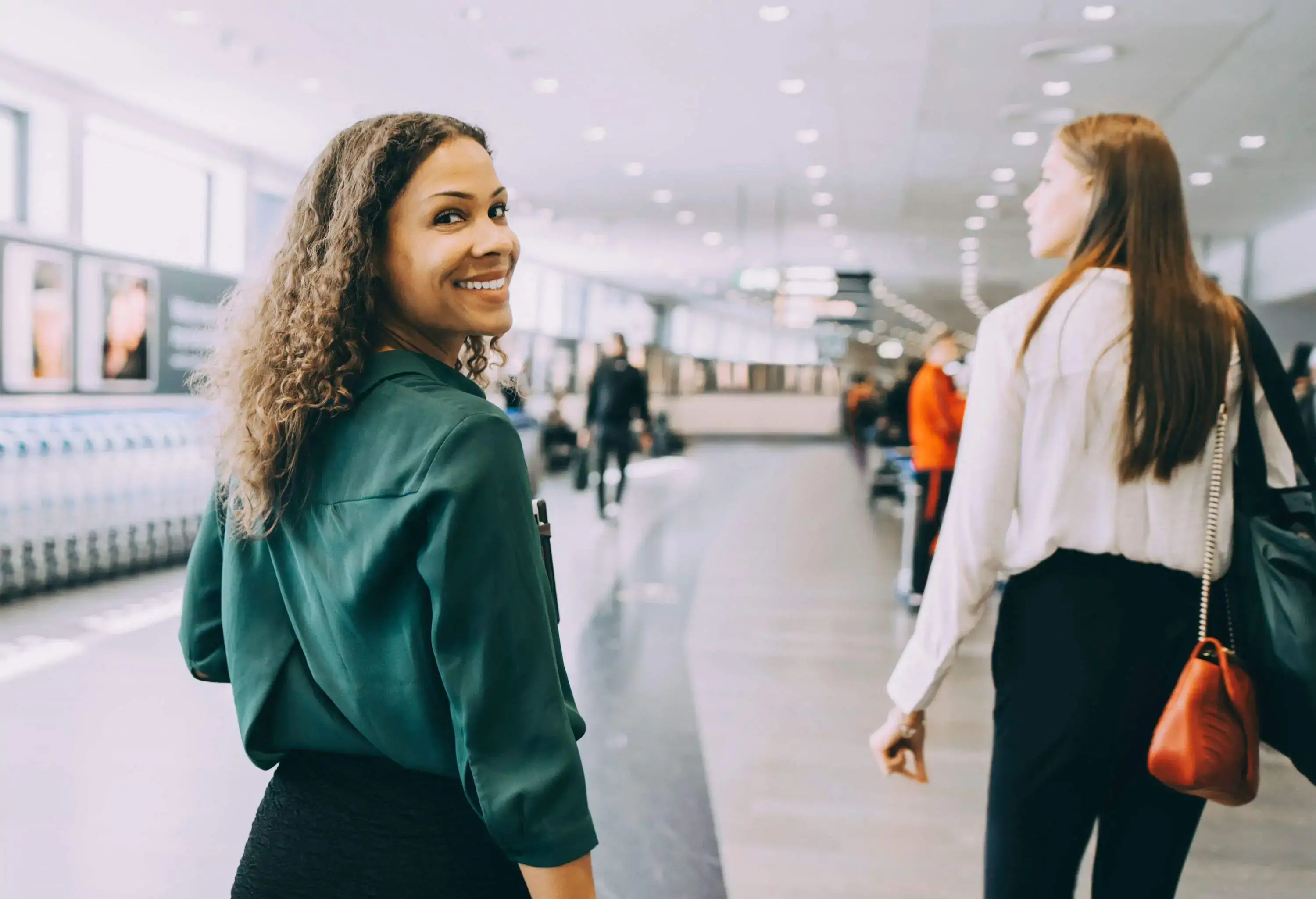 back view of two female colleagues walking through airport, one looking at the camera smiling