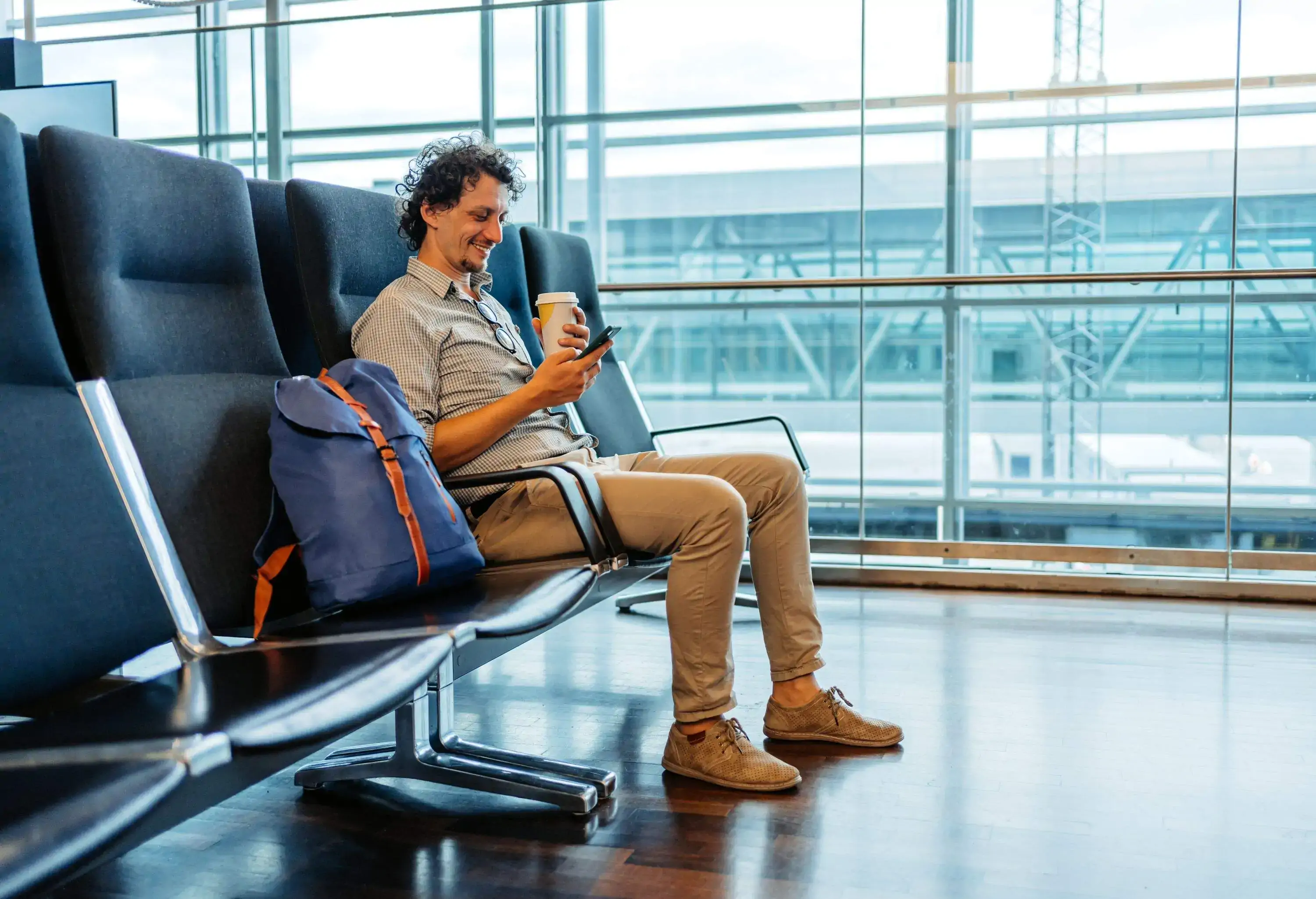 Portrait of smiling man sitting at airport gate holding a take away coffee and a mobile device, backpack standing next to him