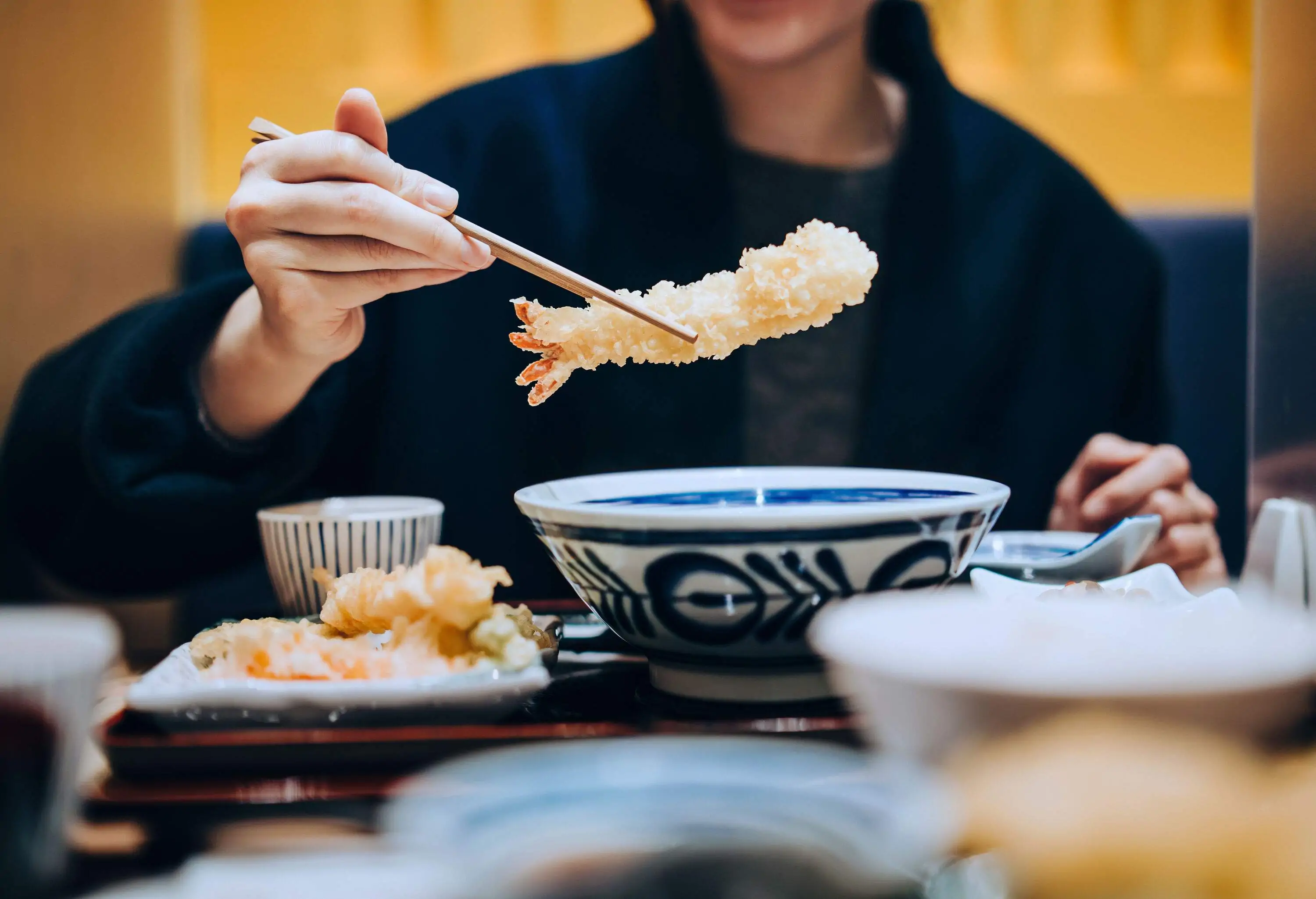 A person holding a piece of tempura using chopsticks, surrounded by a variety of mouth-watering Japanese dishes on the table.