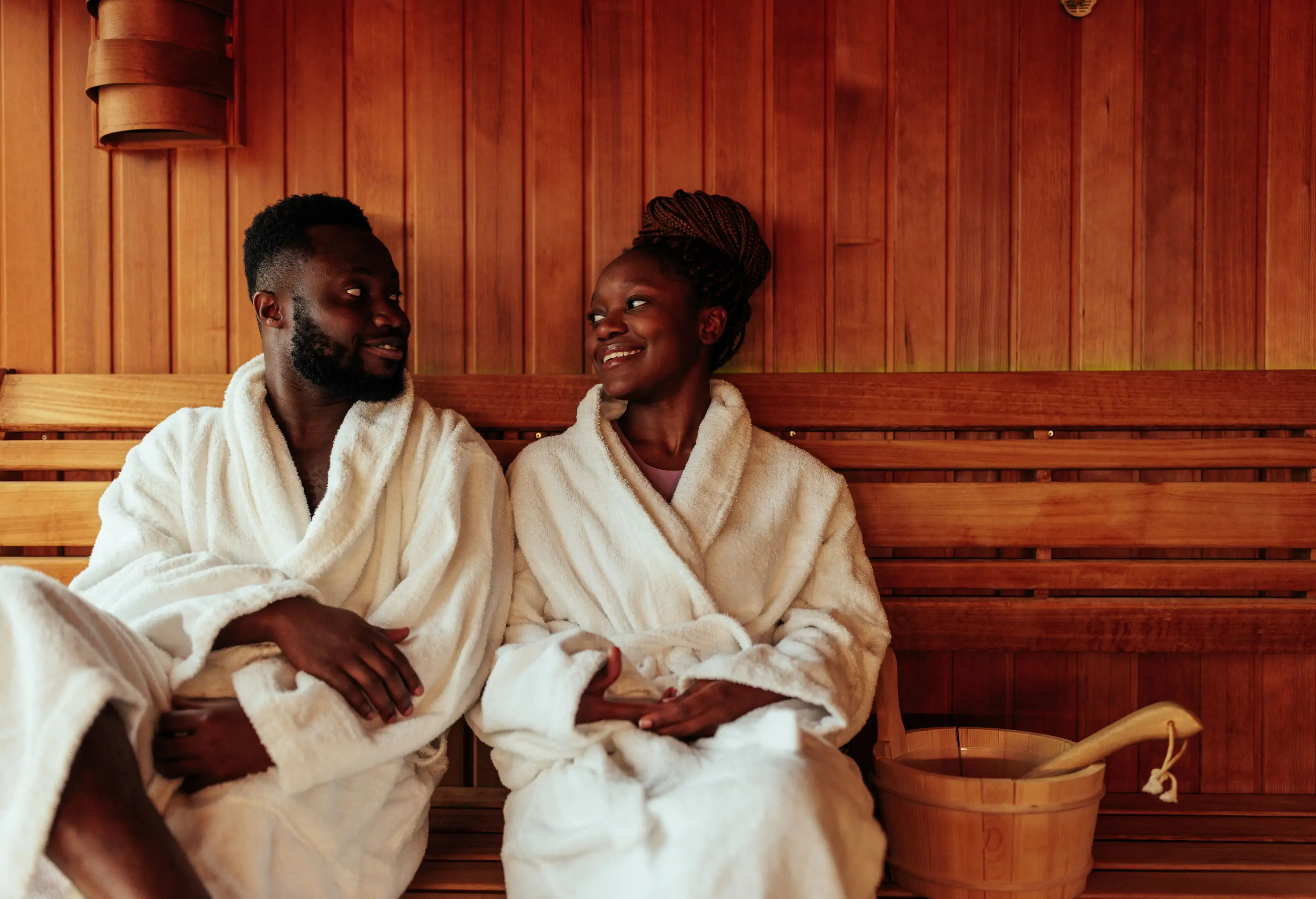 A happy couple is in the sauna. They are wearing bathrobes and are lovingly looking at each other.