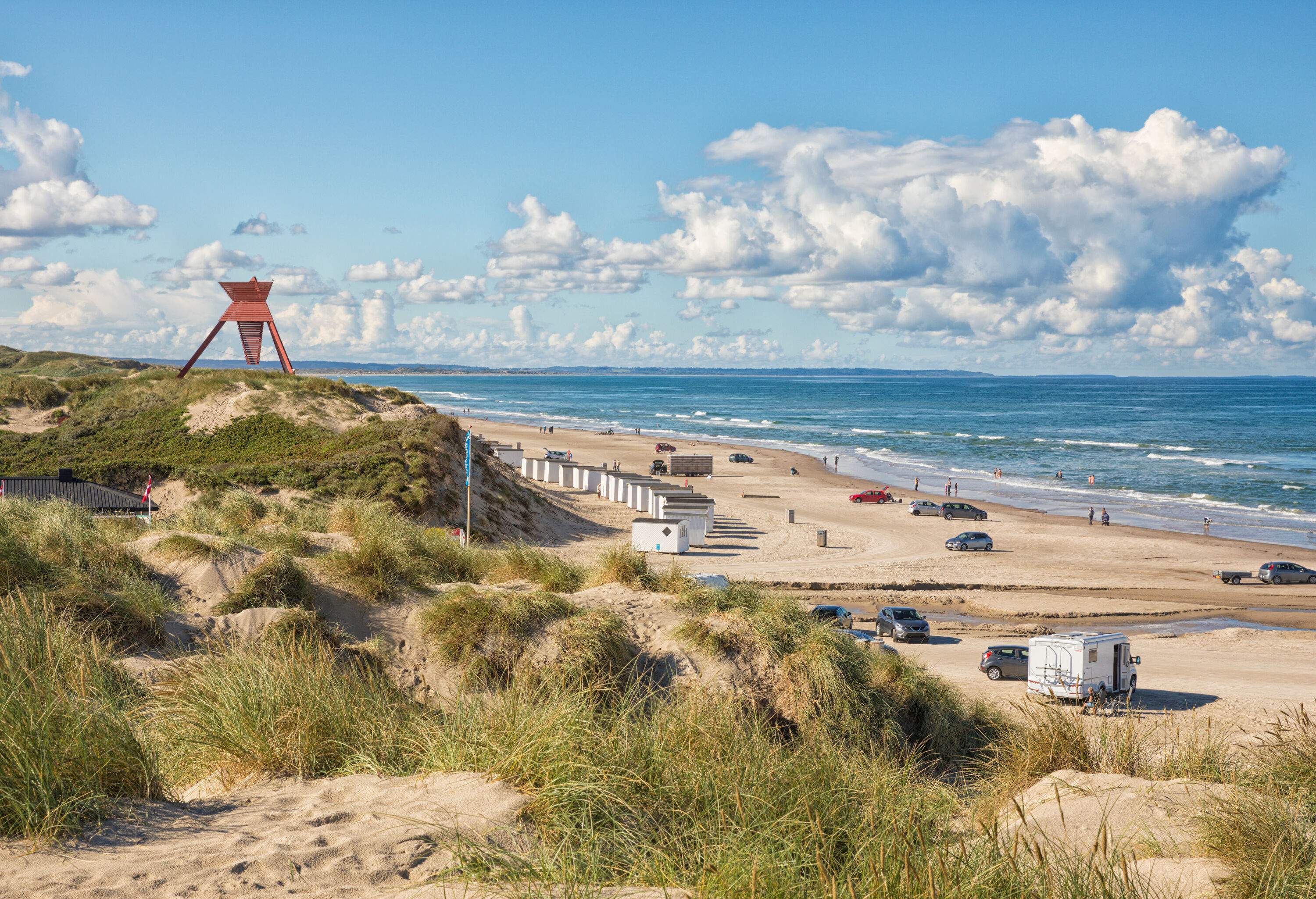 Vehicles parked on the beach, next to beach huts and patches of grass.