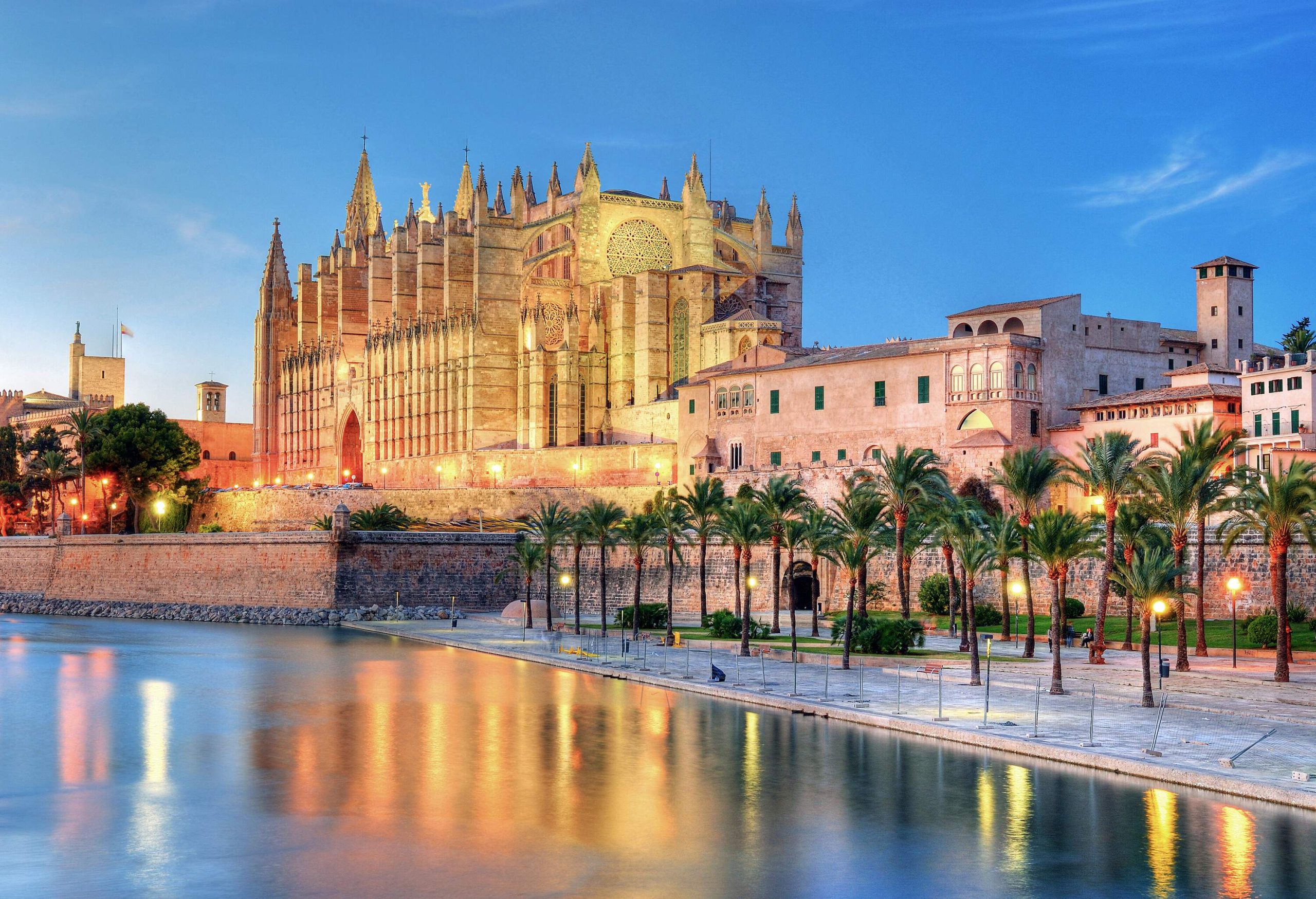 A large mediaeval cathedral with several spires on its top and side walls, surrounded by buildings, can be seen across a tranquil lake.