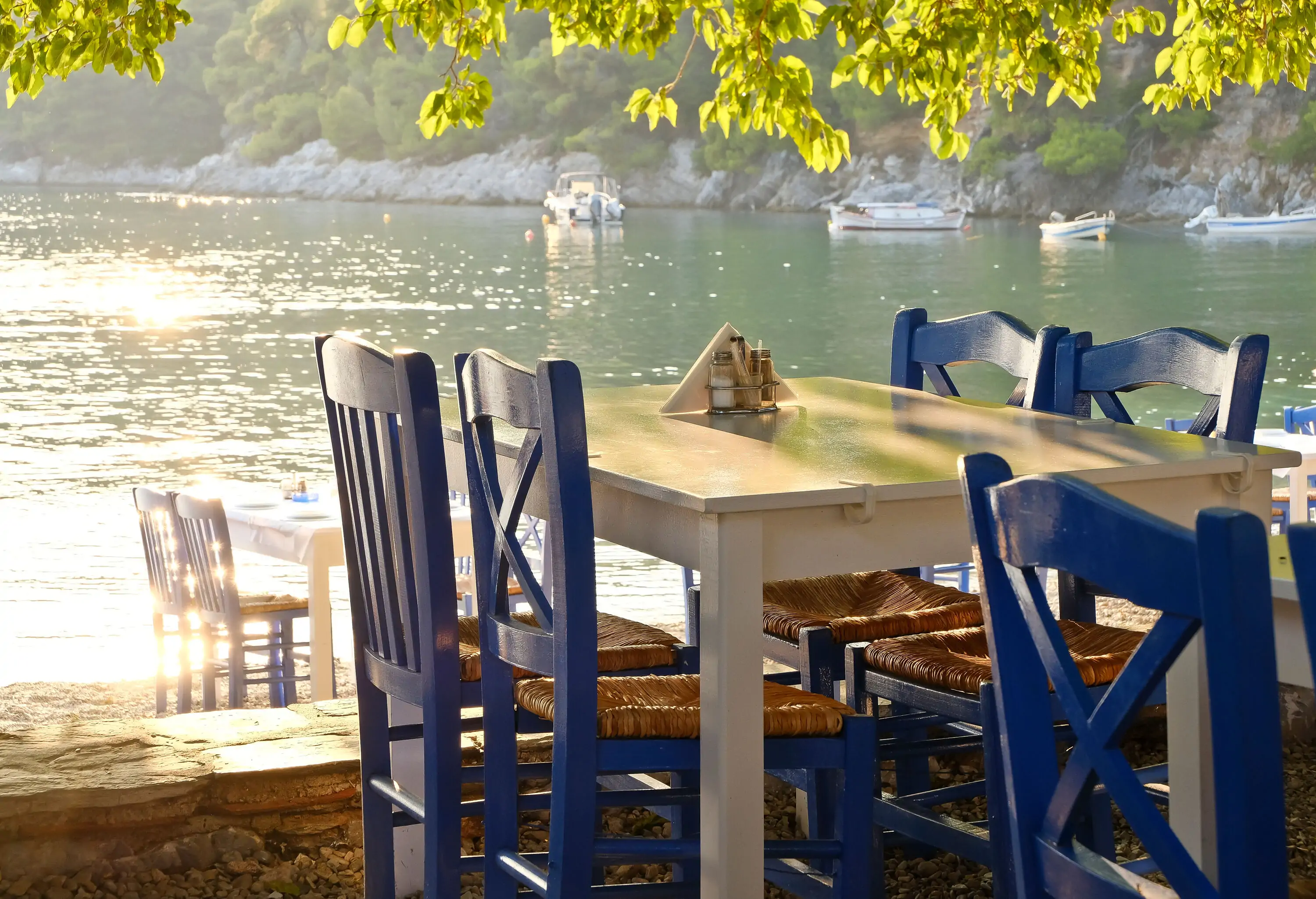 Blue wooden chairs and white tables on the shore of the glimmering sea with moored boats.