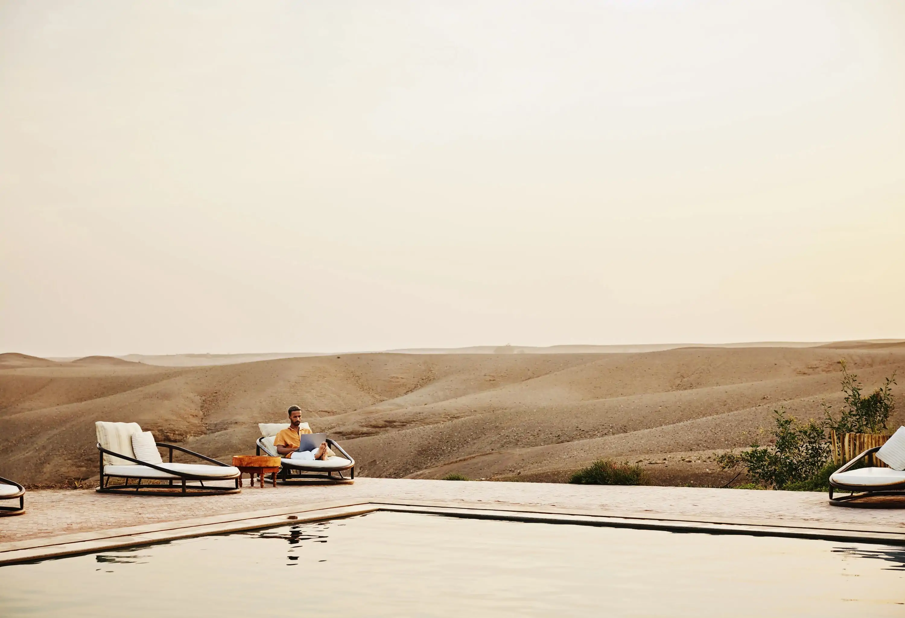 Wide shot of man working on laptop while sitting beside pool at Moroccan desert camp while on vacation