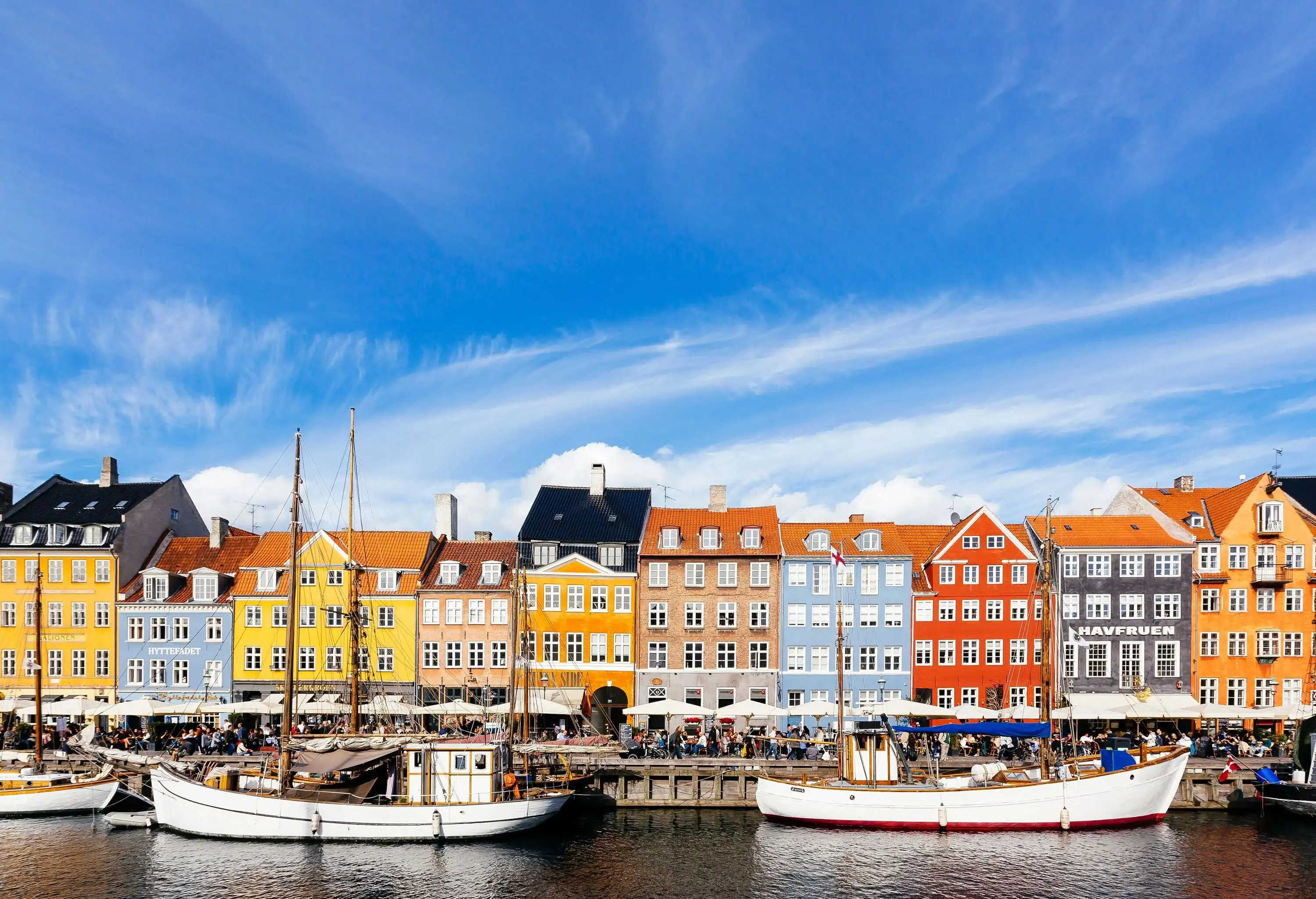 A bustling promenade surrounded by a row of colourful buildings along a harbour with several boats.