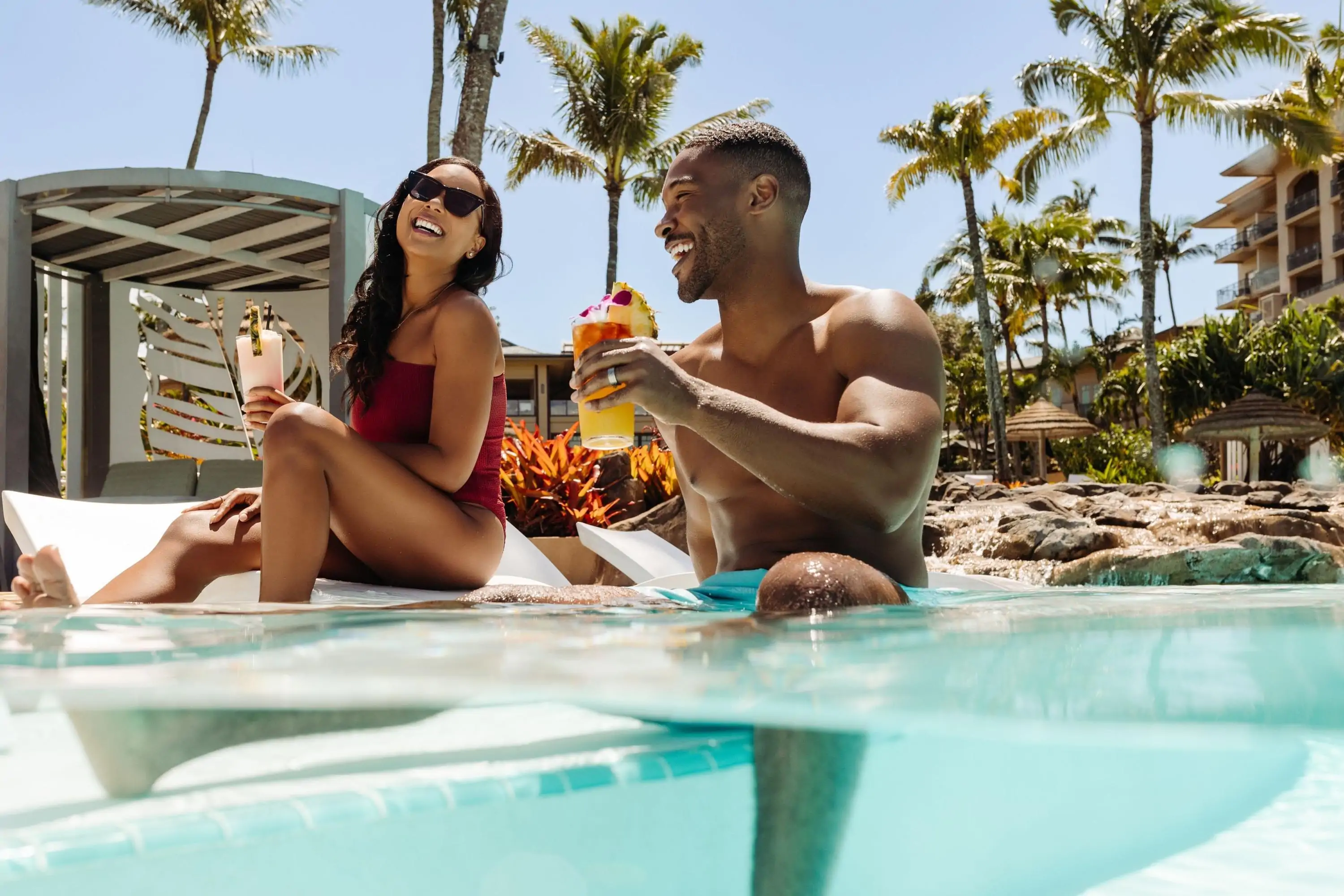 Couple laughing by the pool by a tropical beach laughing and. drinking cocktails