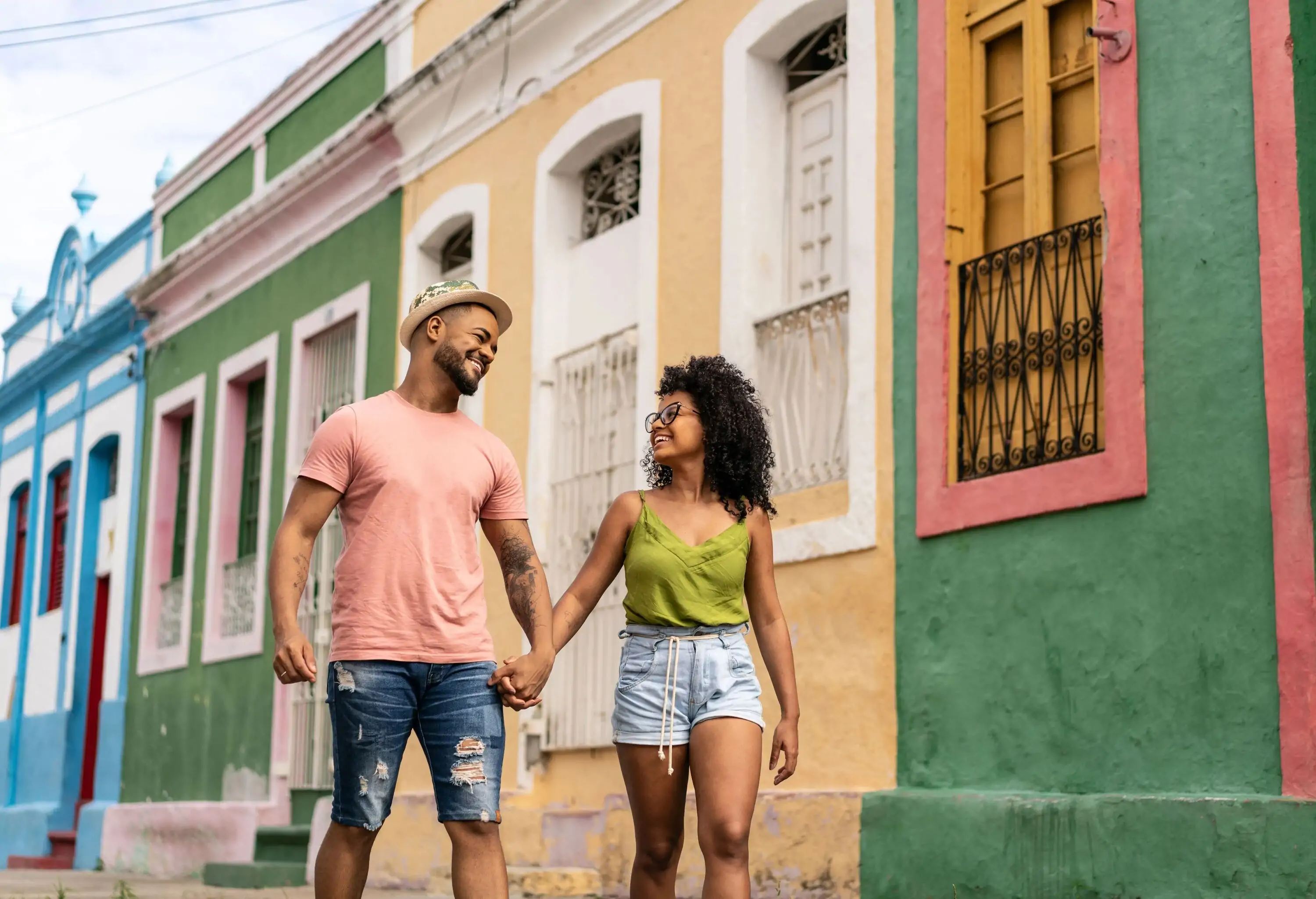 A loving couple walks hand in hand on a charming street, gazing at each other with warm smiles, while a line of colourful stone houses adds to the delightful backdrop.