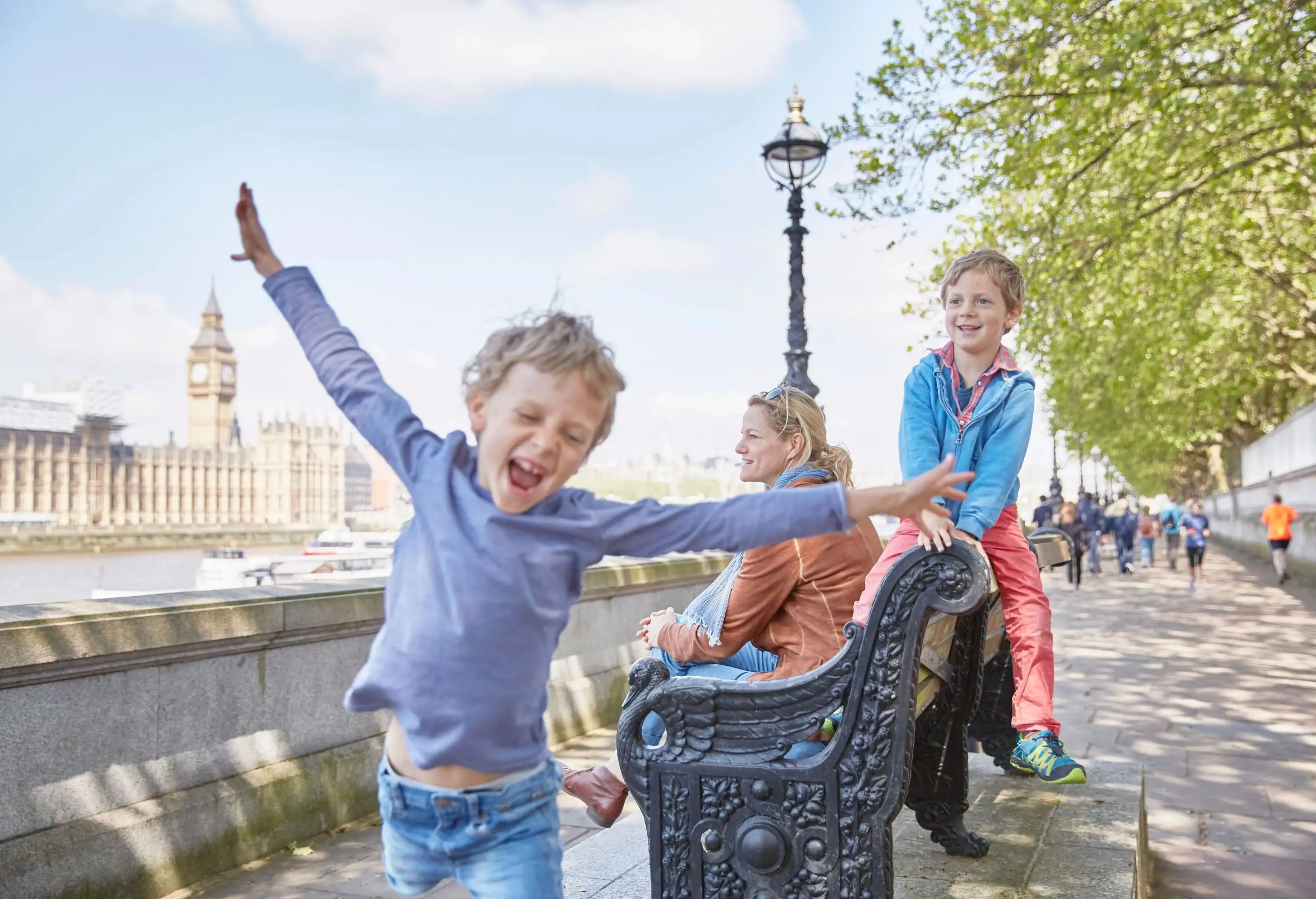 A boy runs with his arms spread out in front of a woman and a child sitting on a sidewalk bench.