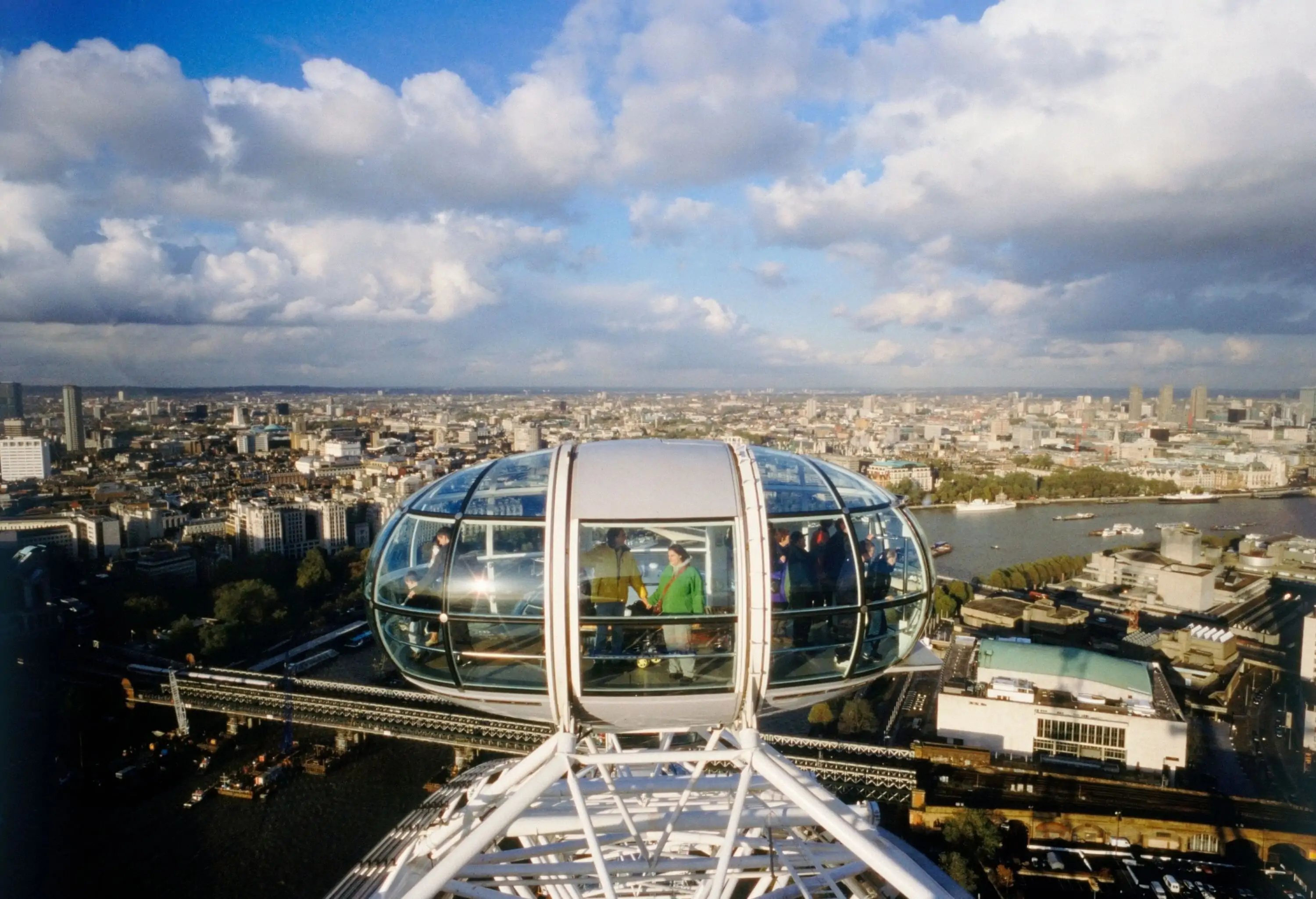 Tourists stand inside a Ferris wheel cabin overlooking a river across a city.