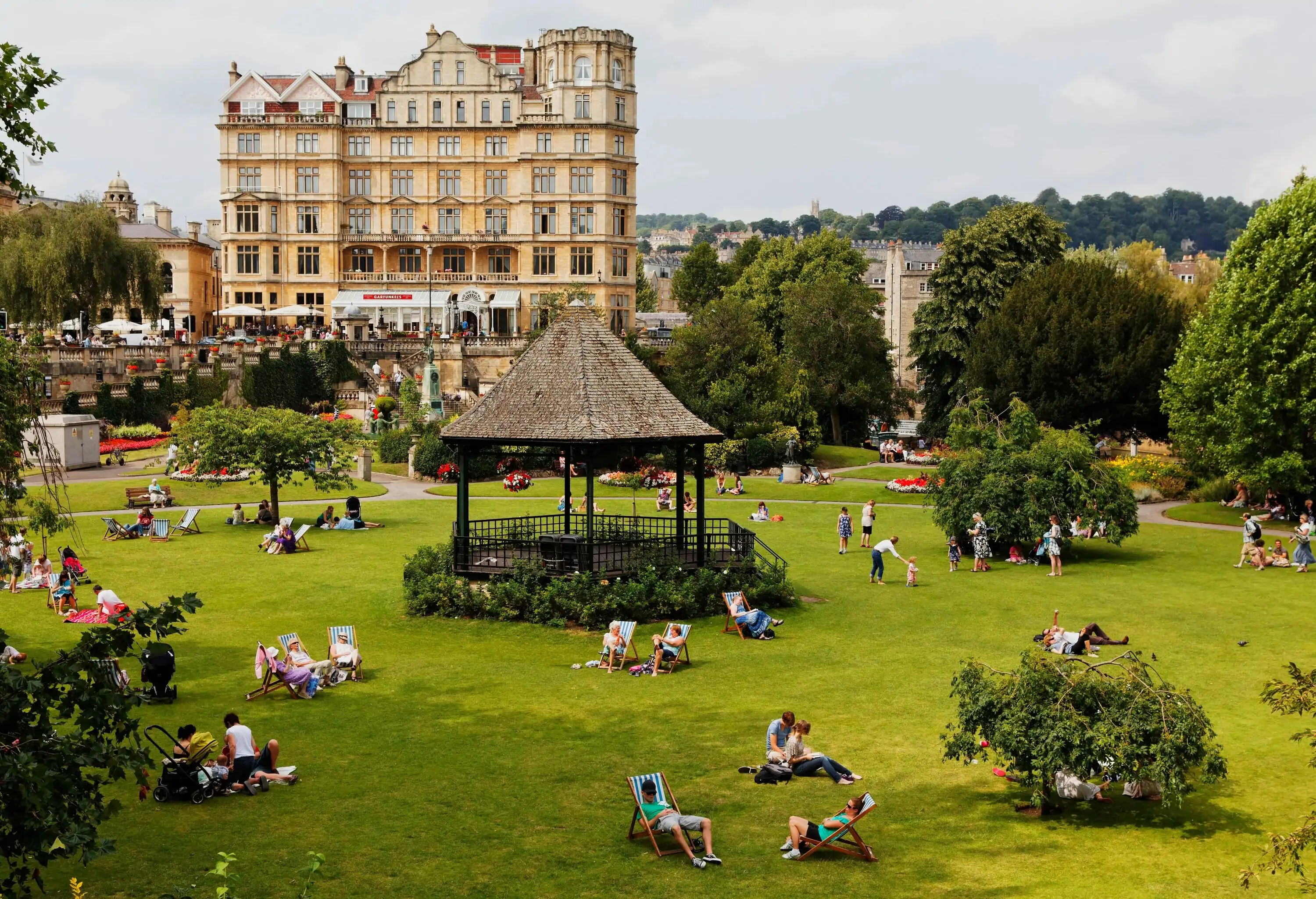 A pavilion in the centre of a lush park full of people overlooking a classic building across.