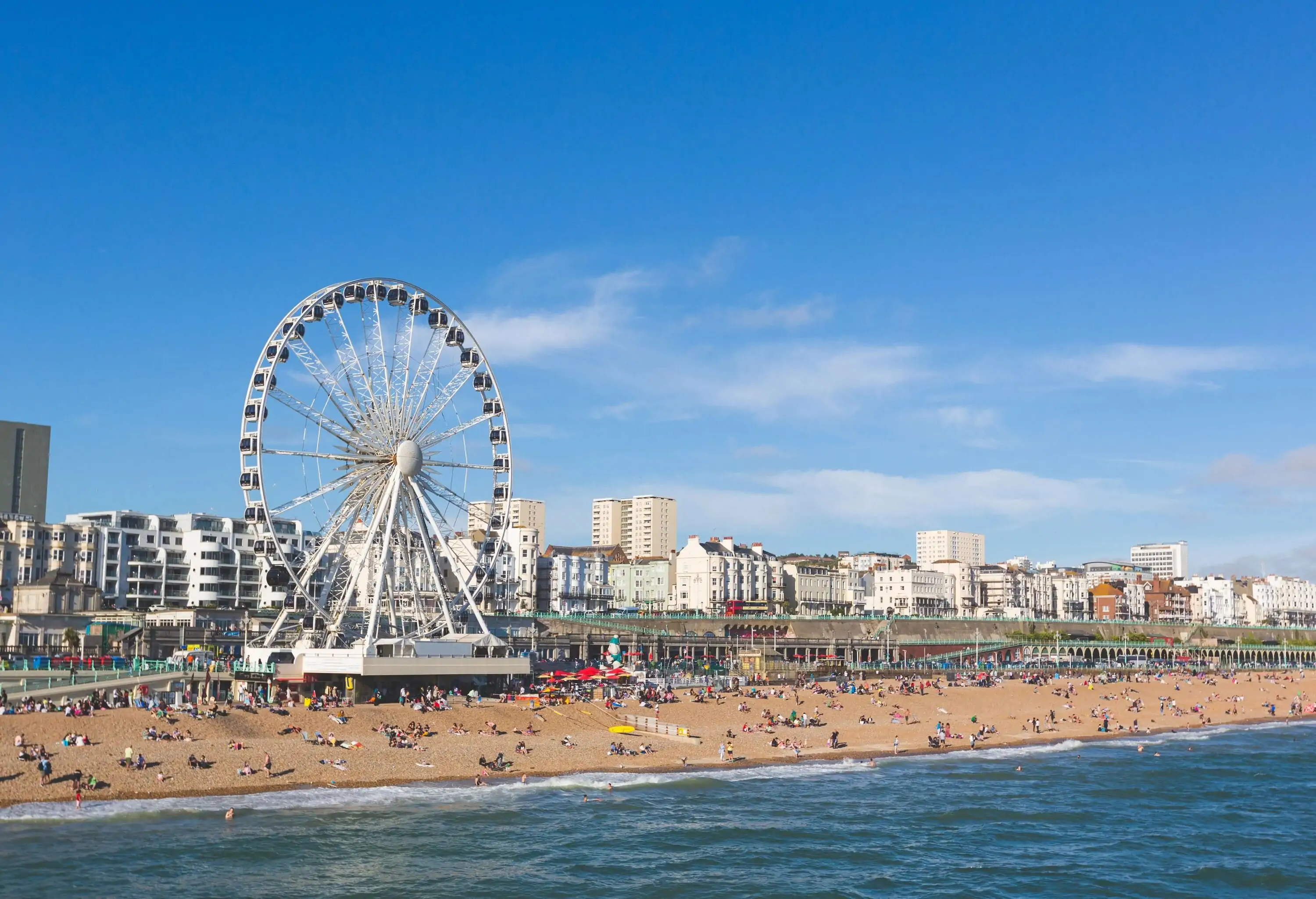 Brighton view of seaside from the pier. Panoramic shot with the famous ferris wheel, the stones beach with unrecognizable persons on a sunny summer day.