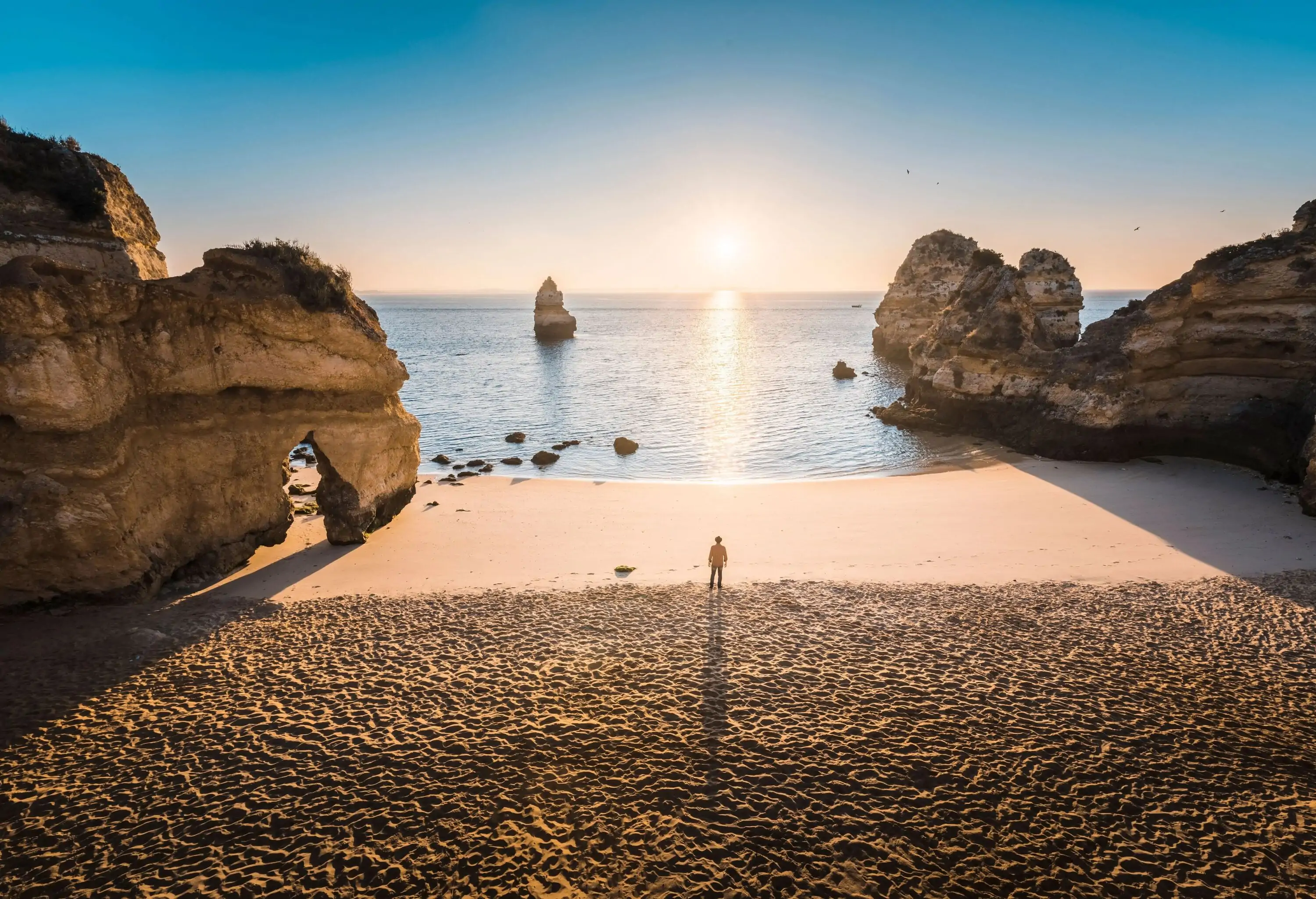 A man is standing between two huge rock formations on the beach at sunrise.