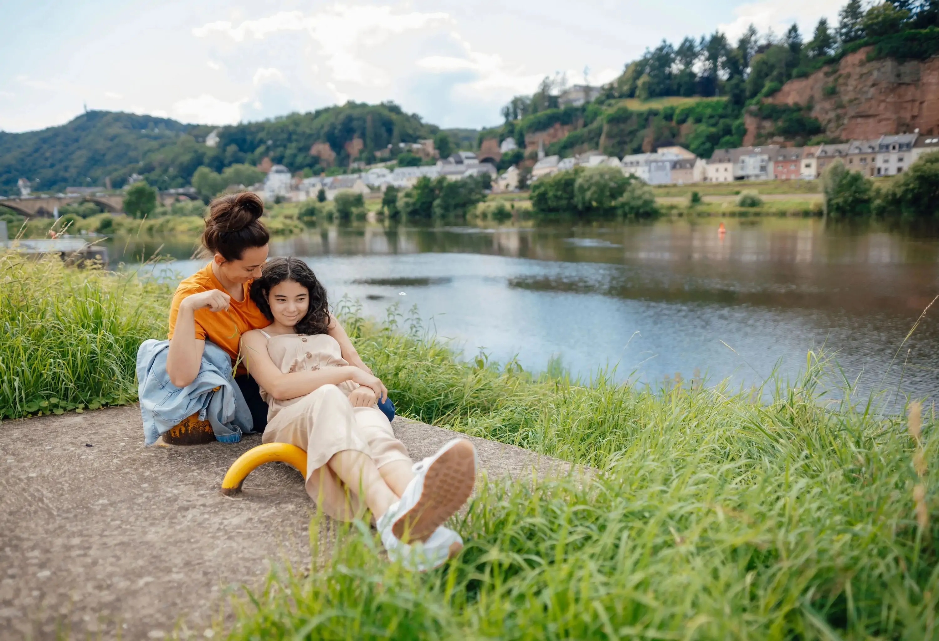 A tender moment shared between a mother and daughter as they sit on a concrete riverbank, the quaint charm of a village visible across the gently flowing water.