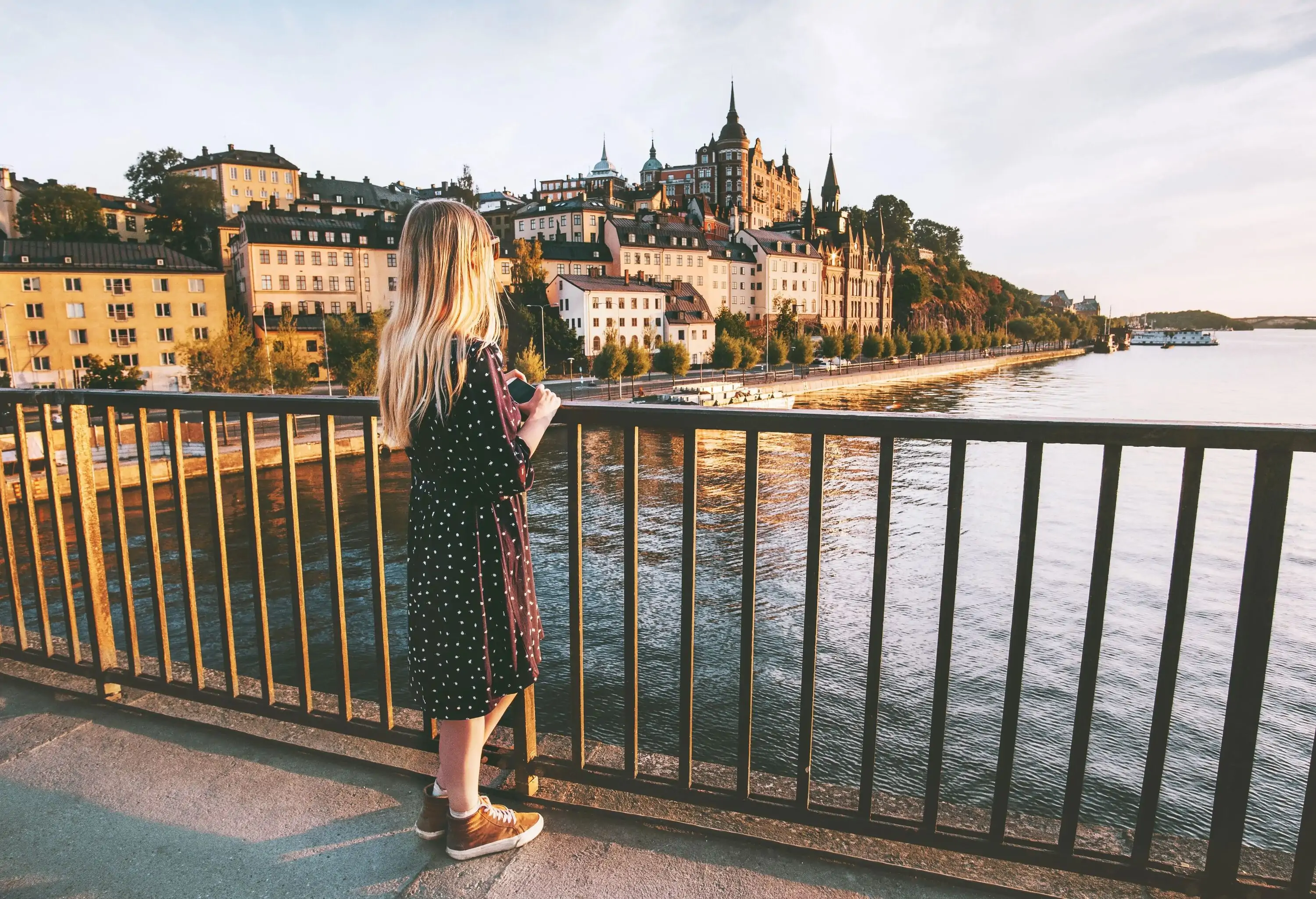 A blonde woman in a black dress leans on the railings of a bridge across a lake and overlooks a cityscape.